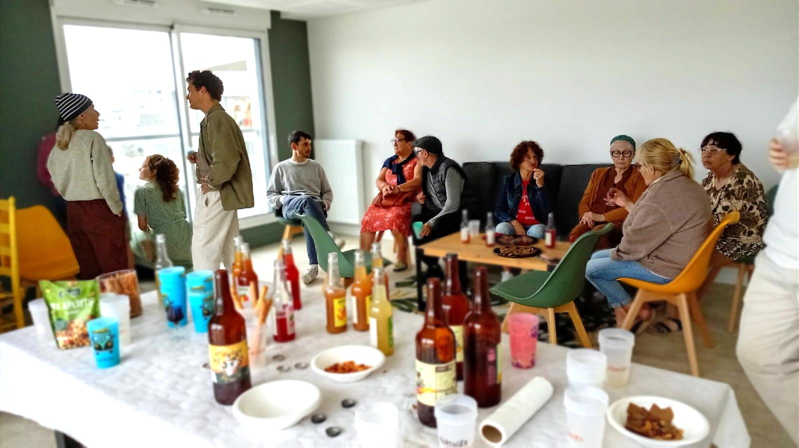 Un groupe de résidents échange autour d’une dégustation de boissons locales dans une salle conviviale, avec plusieurs bouteilles et gobelets disposés sur une table au premier plan.